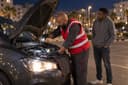 De nuit, un dépanneur inspecte le moteur d’une voiture à la lampe, capot ouvert, pendant que le conducteur attend à côté.
