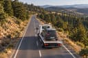 Vue aérienne d’une dépanneuse transportant une voiture sur une route de montagne boisée.