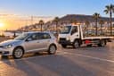 Agadir : une citadine et une dépanneuse vide stationnées face à la marina au coucher du soleil, colline de la Kasbah au loin.